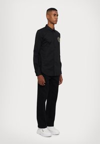 Camicetta nera con bottoni e un logo dorato sul petto, abbinata a pantaloni neri e scarpe da ginnastica bianche. La camicia ha maniche lunghe e un colletto classico.