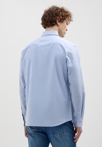 Mann mit lockigem Haar, der ein hellblaues Langarmhemd und blaue Jeans trägt, steht mit dem Rücken zur Kamera vor einem einfarbigen Hintergrund.