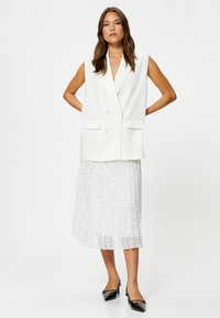 Weißer ärmelloser Blazer mit doppelreihigen Knöpfen, zwei Fronttaschen, kombiniert mit einem fließenden weißen Rock mit schwarzen Punkten und schwarzen High Heels.