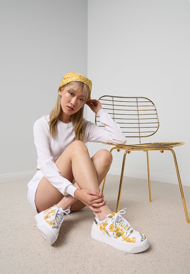 Weiße Sneakers mit gelbem Blumenmuster und schwarzen Akzenten. Modell trägt ein langärmliges weißes Oberteil und sitzt auf einem beigen Teppich neben einem goldenen Stuhl.