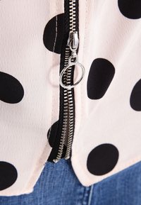 Ljusrosa blus med stora svarta prickar, utrustad med ett silverfärgat dragkedjestängning och texturerad tyg, parat med denim nertill.