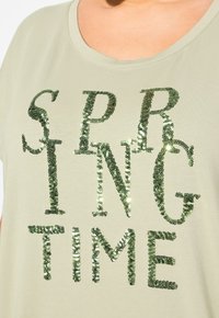 Camiseta verde claro con cuello redondo, que presenta las palabras "TIEMPO DE PRIMAVERA" en grandes y brillantes lentejuelas verdes.