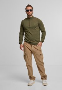 Mann mit Bart und Dutt trägt dunkle Sonnenbrille, olivgrünen Reißverschluss-Pullover, beige Cargo-Hosen und weiße Sneakers und steht vor einem schlichten Hintergrund.