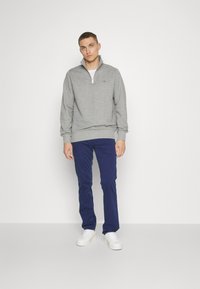 Jeune homme debout portant un pantalon bleu marine, un sweat-shirt gris à fermeture éclair au tiers, des baskets blanches, sur un fond blanc uni.