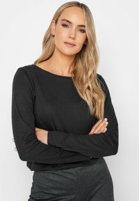Schwarzes Langarmshirt mit rundem Halsausschnitt, aus weichem Stoff. Das Modell steht mit verschränkten Armen und zeigt den entspannten Schnitt und das schlichte Design.