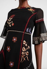 Musta mekko, jossa on kirjailtuja kukkakuvioita punaisena, beigeä ja sinistä. Läpinäkyvät, levenevät hihat koristeellisilla reunoilla ja siksak-ompeleilla.