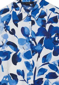 Blue silk blouse featuring a floral design with varying shades of blue leaves on a white background. Classic collar and button closure.