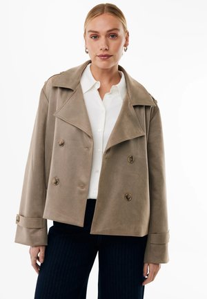 Blonde vrouw met een beige double-breasted jasje over een witte blouse met knopen en donkere broek, staand voor een witte achtergrond.