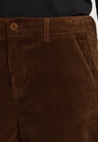 Gros plan sur un pantalon en velours côtelé marron avec un bouton et un passant de ceinture porté avec un haut noir.