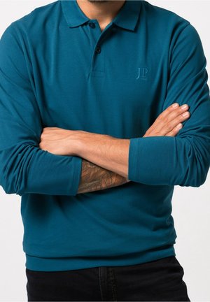 Mand med krydsede arme iført en teal langærmet polo og sorte bukser, der viser en tatovering på sin venstre underarm.