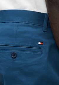 Ανδρικά μπλε chino παντελόνια κατασκευασμένα από απαλό ύφασμα, με τσεπάκι στην πλάτη με κουμπί και ένα μικρό λογότυπο σε κόκκινο, λευκό και μαύρο.