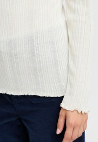 Suéter de punto blanco con canalé y mangas largas, con un dobladillo y puños con forma ondulada. La tela tiene un aspecto suave y texturizado.