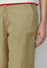Pantaloni beige con una texture liscia, vestibilità sartoriale e tasche laterali; cuciture a contrasto e una mano posizionata sulla vita.