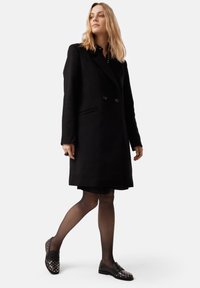 Manteau en laine noir avec fermeture double-breasted, poches latérales et coupe ajustée, porté par-dessus une robe noire et accessoirisé avec des chaussures à clous.