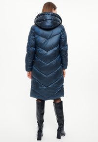 Donkerblauwe pufferjack met een chevronpatroon en een grote capuchon. Het materiaal is glanzend en het heeft lange mouwen en is tot de knie. Zwarte kniehoogte laarzen zijn zichtbaar.