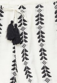 Tissu blanc avec des motifs géométriques verticaux noirs et un cordon de serrage noué orné de deux pompons noirs.