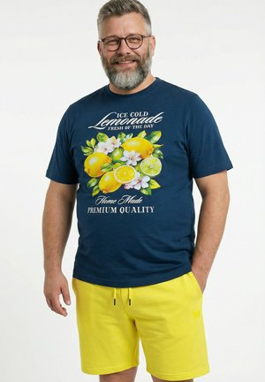 Uśmiechnięty mężczyzna w okularach nosi granatową koszulkę z nadrukiem cytryny i kwiatu oraz napisem „Ice Cold Lemonade”, w połączeniu z jaskrawożółtymi szortami.