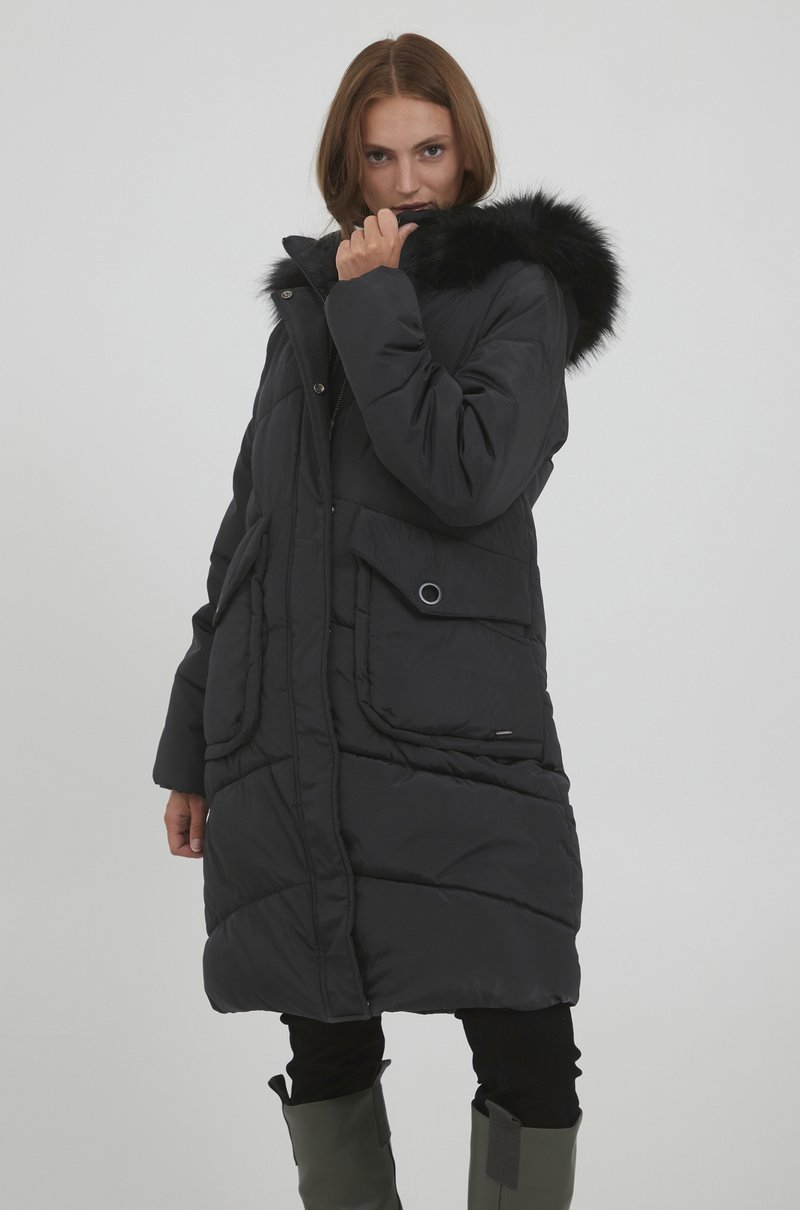 Sort dunjakke med en faux fur hætte, der har et quiltet mønster, store forlommer og lynlås, båret over sorte bukser.