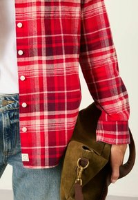 Red and white plaid flannel shirt with buttons, worn over a white t-shirt and paired with blue jeans and a brown suede bag.