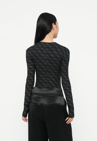 Donna con lunghi capelli neri che indossa un top lungo a maniche fitte con motivo neri e pantaloni larghi in satin nero, di spalle contro uno sfondo bianco.