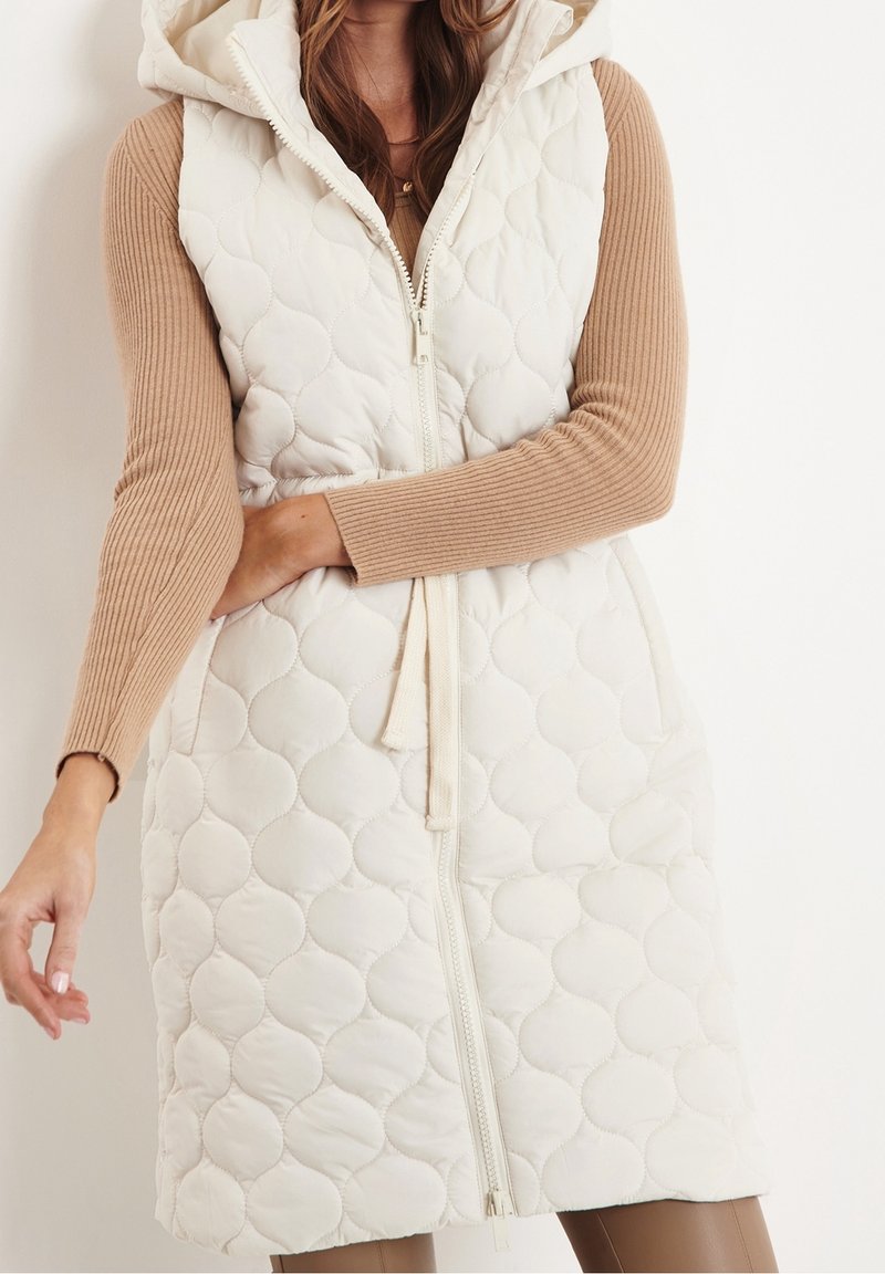 Femme portant une veste matelassée blanche sans manches à capuche, par-dessus un haut beige à manches longues côtelées, debout devant un fond clair uni.