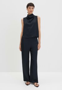 Top nero senza maniche con colletto drappeggiato abbinato a pantaloni a gamba larga. Il tessuto ha una texture liscia e lucida. Design e silhouette minimalistici.