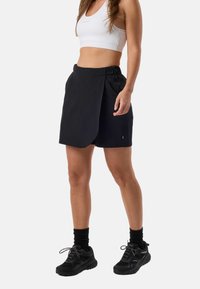 Kvinde iført en hvid sports-BH, sort omslagsnederdel med shorts, sorte sokker og sorte sportssko, stående foran en ensfarvet baggrund.