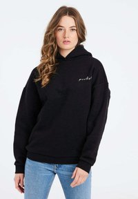 Felpa nera con cappuccio e maniche lunghe, con tasca frontale e logo bianco ricamato. Abbinata a jeans azzurri chiari.