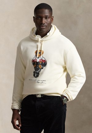 Homme portant un sweat à capuche crème avec un motif Polo Bear et un pantalon noir, debout devant un fond beige uni.