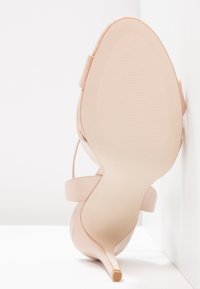Sandale beige à talon haut vue de la semelle, avec un talon fin et une bride à la cheville sur fond blanc.