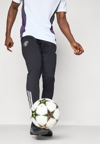 Svart treningsbukser med en logo, kombinert med en hvit og lilla T-skjorte. En mønstrete hvit fotball med grønne detaljer ligger på bakken.