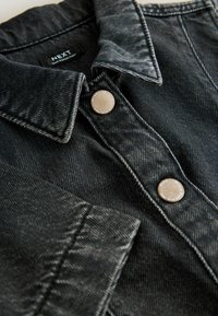 Svart denimjacka med spetsig krage, med två ljusa knappar och kontrasterande sömmar. Strukturerat tyg, närbild.