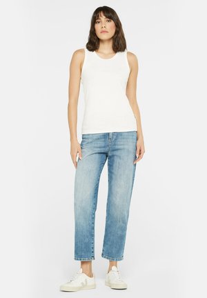 Weißes ärmelloses Oberteil mit rundem Halsausschnitt, kombiniert mit hellblauen hochgeschnittenen Jeans und weißen Sneakern mit subtilen Logo an der Seite.