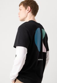 Sort bomulds t-shirt med abstrakt grafik i teal, grøn og pink på bagsiden. Lang hvid ærme lag synlig nedenunder.