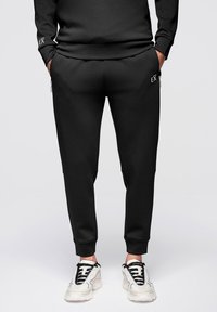 Pantalon de jogging noir avec poches à fermeture éclair porté par une personne en baskets blanches et noires, debout les mains dans les poches.