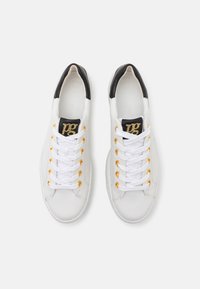 Baskets en cuir blanc avec œillets dorés, talon noir, et logo décoratif en texte doré sur une languette noire. Semelle en caoutchouc, finition texturée.