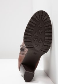 Botte en cuir marron avec semelle noire crantée, posée sur une surface blanche sur fond blanc, talon incliné vers la caméra.