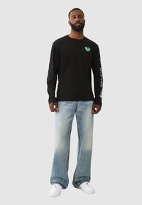 Schwarzes Langarmshirt mit Brustgrafik, hellblaue weit geschnittene Jeans und weiße Sneakers. Mit kontrastierendem Nähen und einem lockeren Schnitt.