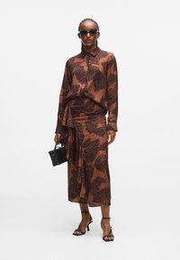 Brązowa sukienka w wzory z pasującą bluzką, z motywem liści. Dodatki to czarna torebka i sandały na wysokim obcasie z paskami.