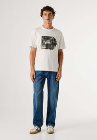 Jonge man in een wit graphic t-shirt, blauwe jeans en witte sneakers, staand tegen een effen lichte achtergrond.