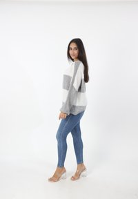 Oversized gebreide trui met grijze en witte strepen. Gecombineerd met een blauwe skinny jeans en transparante blokhakken op een effen achtergrond.