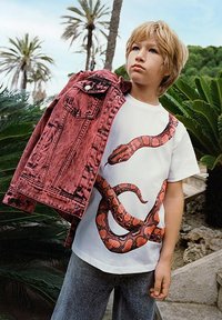 Giacca di jeans rossa lavata acido, tenuta in alto accanto a una maglietta bianca con una grande grafica di un serpente. Indossata con jeans di denim grigio.
