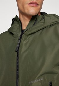 Grüne wasserdichte Jacke mit einer strukturierten Kapuze, Front-Reißverschluss und schwarzen Akzenten. Bietet eine glatte Textur und dezente Markierung am Saum.