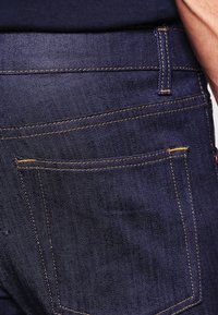 Mörka denimjeans har en närbild av bakfickan med orange sömnad. Tyget har en slät textur och en djup indigofärg.