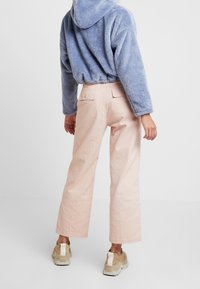 Osoba oblečená v lehce růžových širokých kalhotách, béžových masivních teniskách a světle modré huňaté mikině, stojící před jednobarevným bílým pozadím.