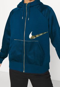 Person klädd i en blå zip-up hoodie med en metallisk guld Nike-logga på bröstet och svarta byxor, stående mot en enfärgad bakgrund.