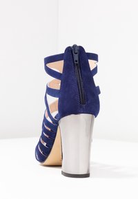 Mörkblå mocka högklackad sko med utskurna remmar och en silverfärgad metallic klack. Baktill med dragkedja som stängning.
