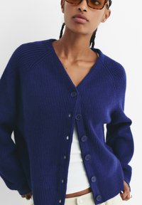 Mornarica rebrasta kardigan s V-izrezom, pet dugmadi na zatvaranju i dugim rukavima. Mekana tekstura i opušteni kroj. Nosiva preko bijelog topa.