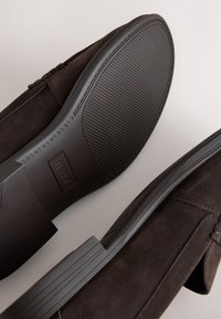 Mocassins en daim marron avec pampilles, montrant une semelle en caoutchouc texturée et un talon embossé du nom de la marque "NEXT".