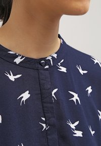 Blouse bleu marine avec motif d'oiseaux blancs, encolure ronde et fermeture à boutons. Tissu lisse au toucher doux.